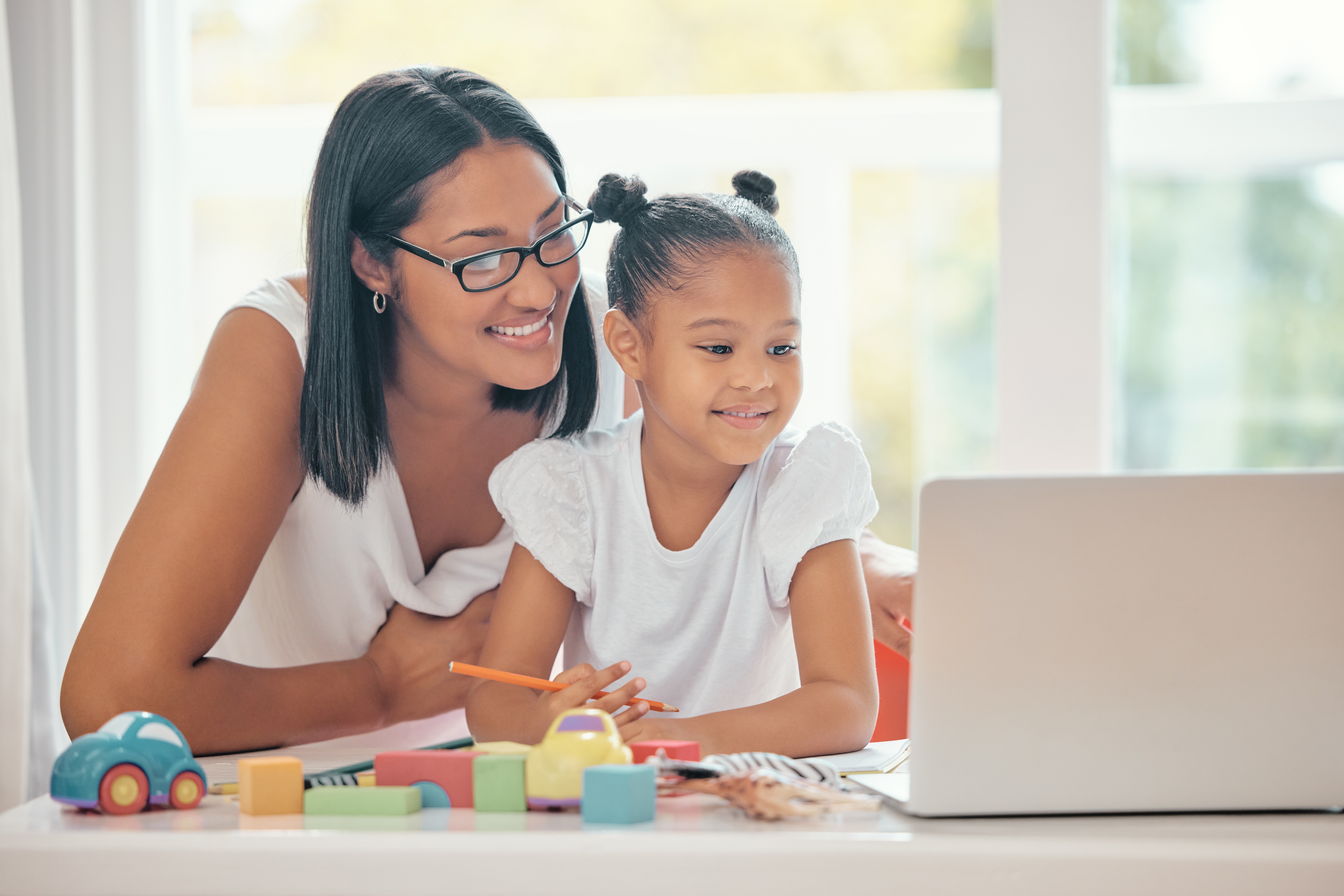 A mom and her young daughter smile at a laptop they are using for homeschooling, with school supplies in front of them.